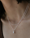 A close-up image of a person's neck adorned with two delicate necklaces from Becoming Jewelry. The lower Shine Necklace features a small pendant accented by a sparkling charm.
