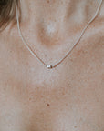 A person sporting a Becoming Jewelry Stick Together Necklace, featuring a small pendant on a silver chain, over a gray top, symbolizing a special connection.