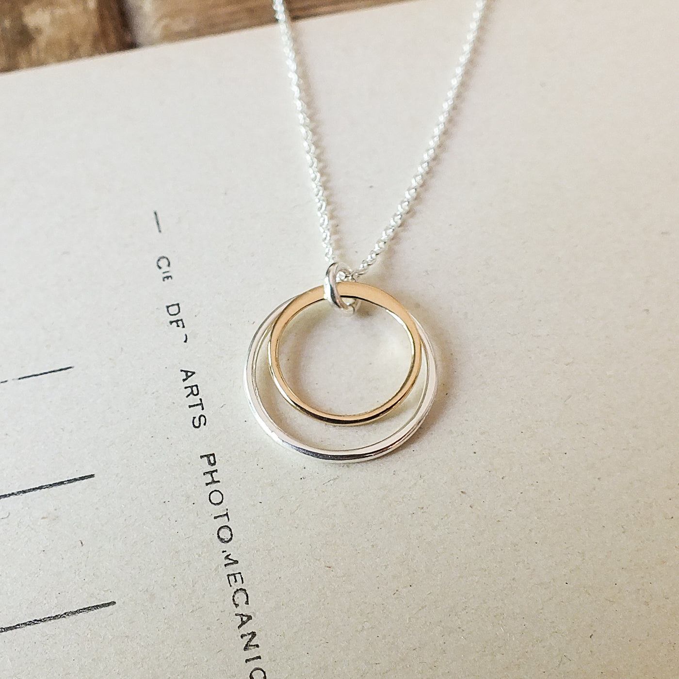 A Surrounded By Love Necklace by Becoming Jewelry with a pendant of three interlinked mixed metal rings, silver and gold, resting on an open book.