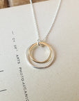 The Surrounded By Love Necklace by Becoming Jewelry features a silver pendant with two interlocking rings, one gold and one silver, elegantly placed on a textured beige surface. This refined piece makes the perfect gift for someone special and serves as a meaningful reminder of your bond.