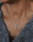A close-up of a person wearing a delicate gold chain necklace with a vertical bar pendant set with small clear stones, paired with a fuzzy gray knit top.