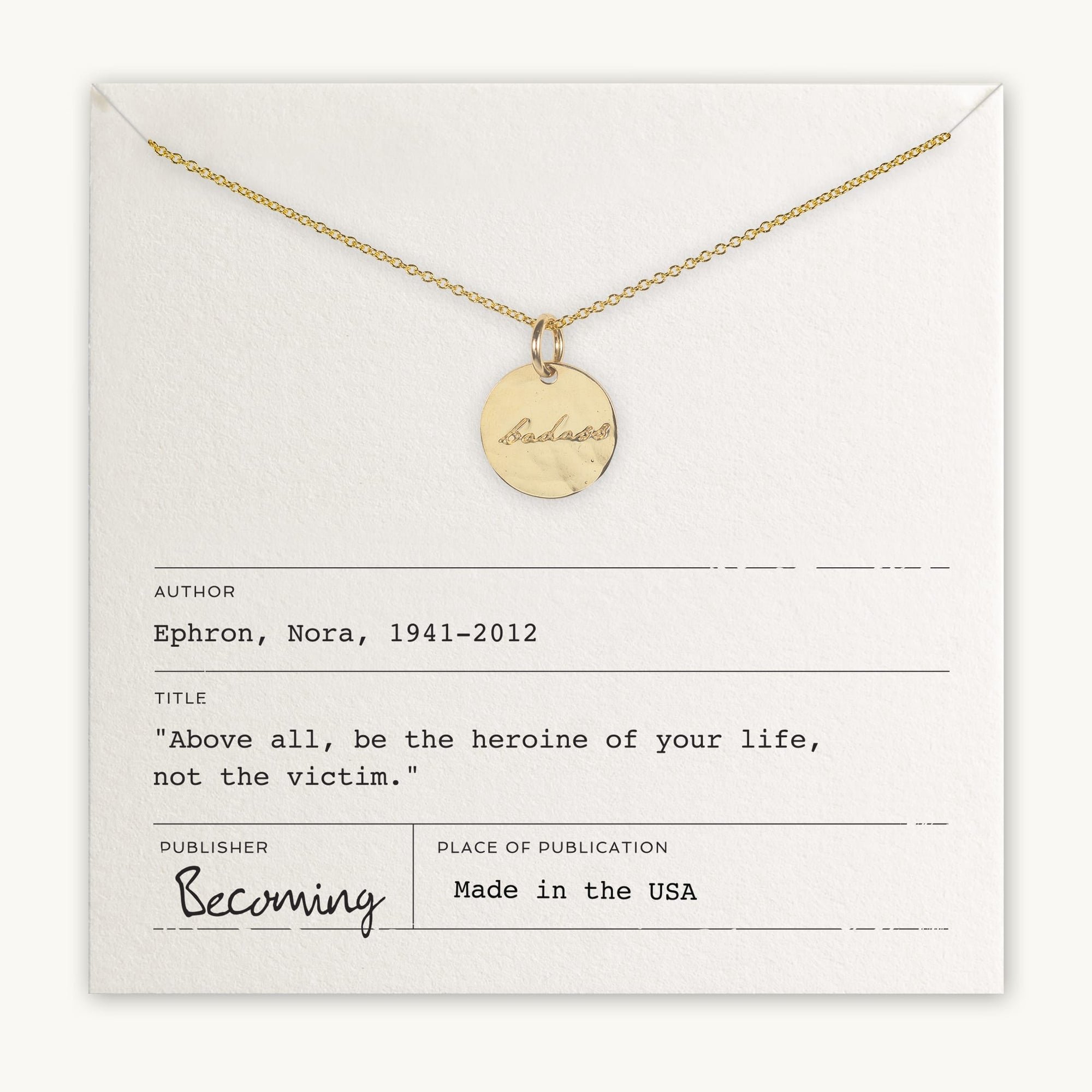 The Badass Necklace from Becoming Jewelry features a gold circular pendant with the word "badass" engraved in an antique handwriting font. It is beautifully displayed on a card that includes a powerful quote by Nora Ephron: "Above all, be the heroine of your life, not the victim." This piece offers you a daily dose of empowerment.