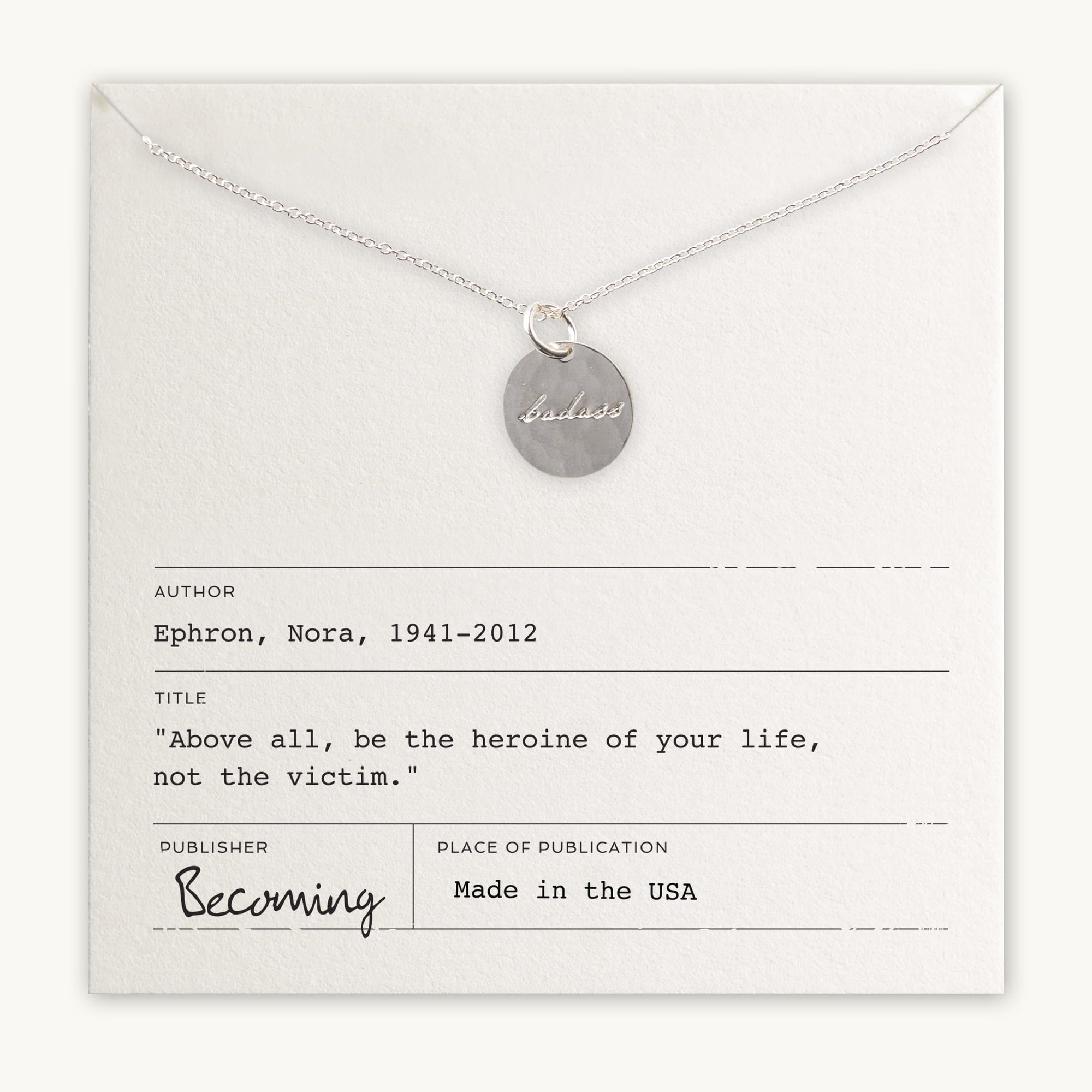 The "Badass Necklace" by Becoming Jewelry showcases a silver necklace with a round pendant inscribed with "badass," elegantly displayed on a card adorned with an antique handwriting font and featuring an inspiring quote by Nora Ephron: "Above all, be the heroine of your life, not the victim." This piece exudes empowerment and elegance.