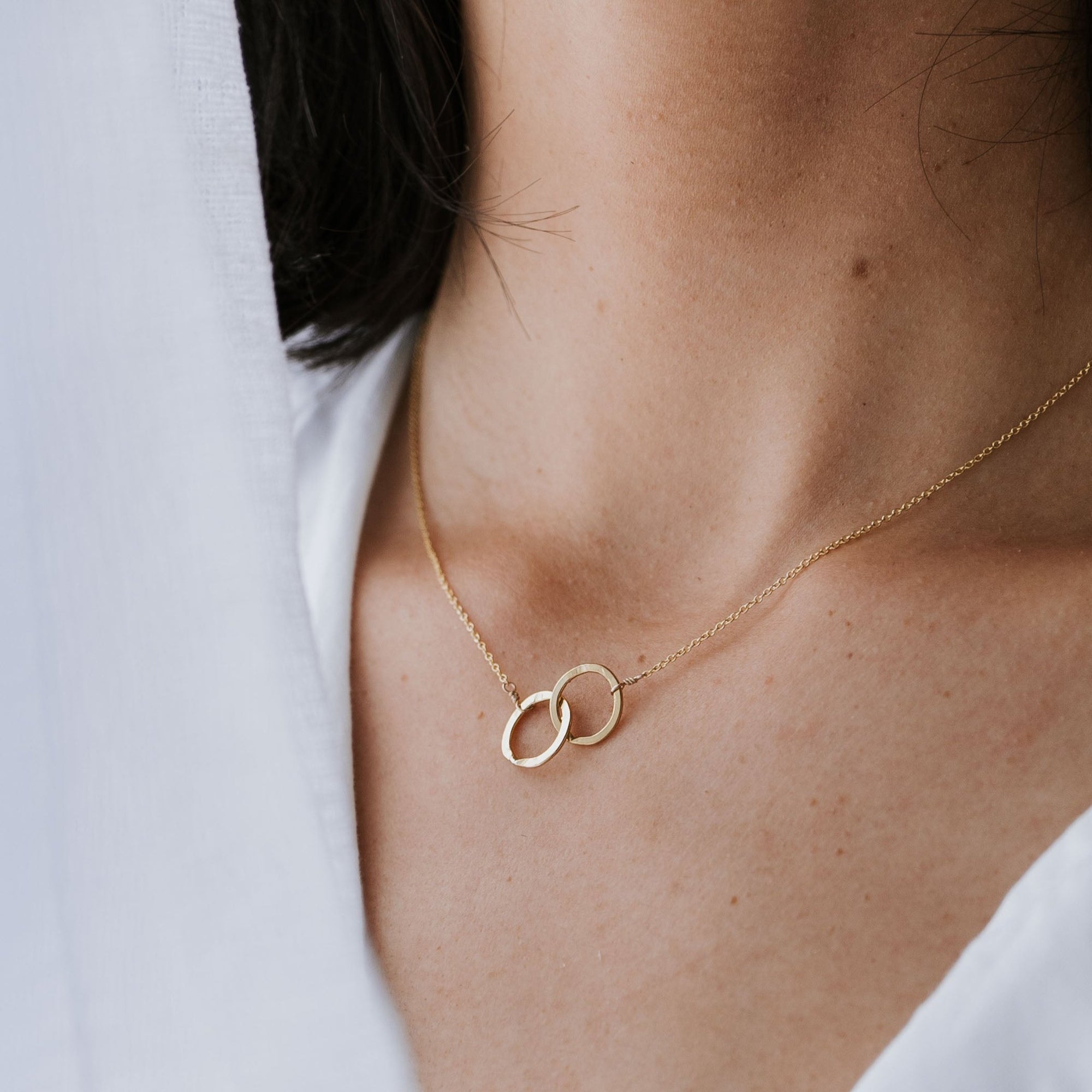 Close-up of a person wearing a delicate gold Joined for Life Necklace from Becoming Jewelry, featuring two interlocking rings that symbolize an unbreakable bond. The background is a soft white fabric, accentuating the elegance of the intertwined circles.