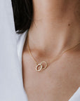 Close-up of a person wearing a delicate gold Joined for Life Necklace from Becoming Jewelry, featuring two interlocking rings that symbolize an unbreakable bond. The background is a soft white fabric, accentuating the elegance of the intertwined circles.