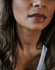 A woman in a white tank top layers her wavy hair over her shoulders, accenting a glossed smile with hoop earrings and the delicate Bead Chain Necklace from Becoming Jewelry.