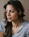 A woman with long hair in a gray shirt looks to the side, her neck adorned by the elegant Figaro Chain Necklace from Becoming Jewelry. The neutral background enhances her serene elegance.