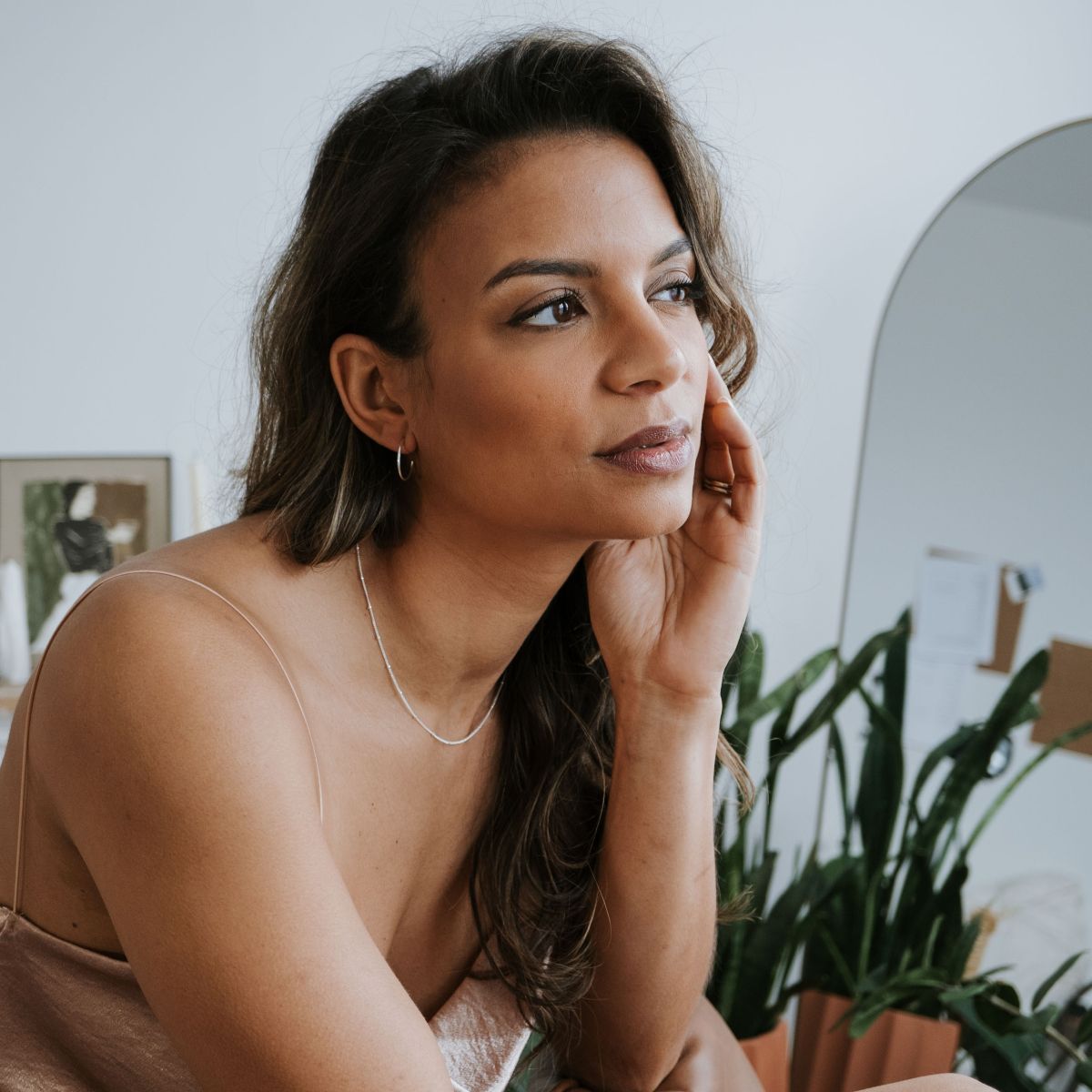 A woman with long hair, seated indoors near plants and a mirror, rests her head on her hand. She wears the Becoming Jewelry Satellite Chain Necklace, which adds elegance to her contemplative gaze.