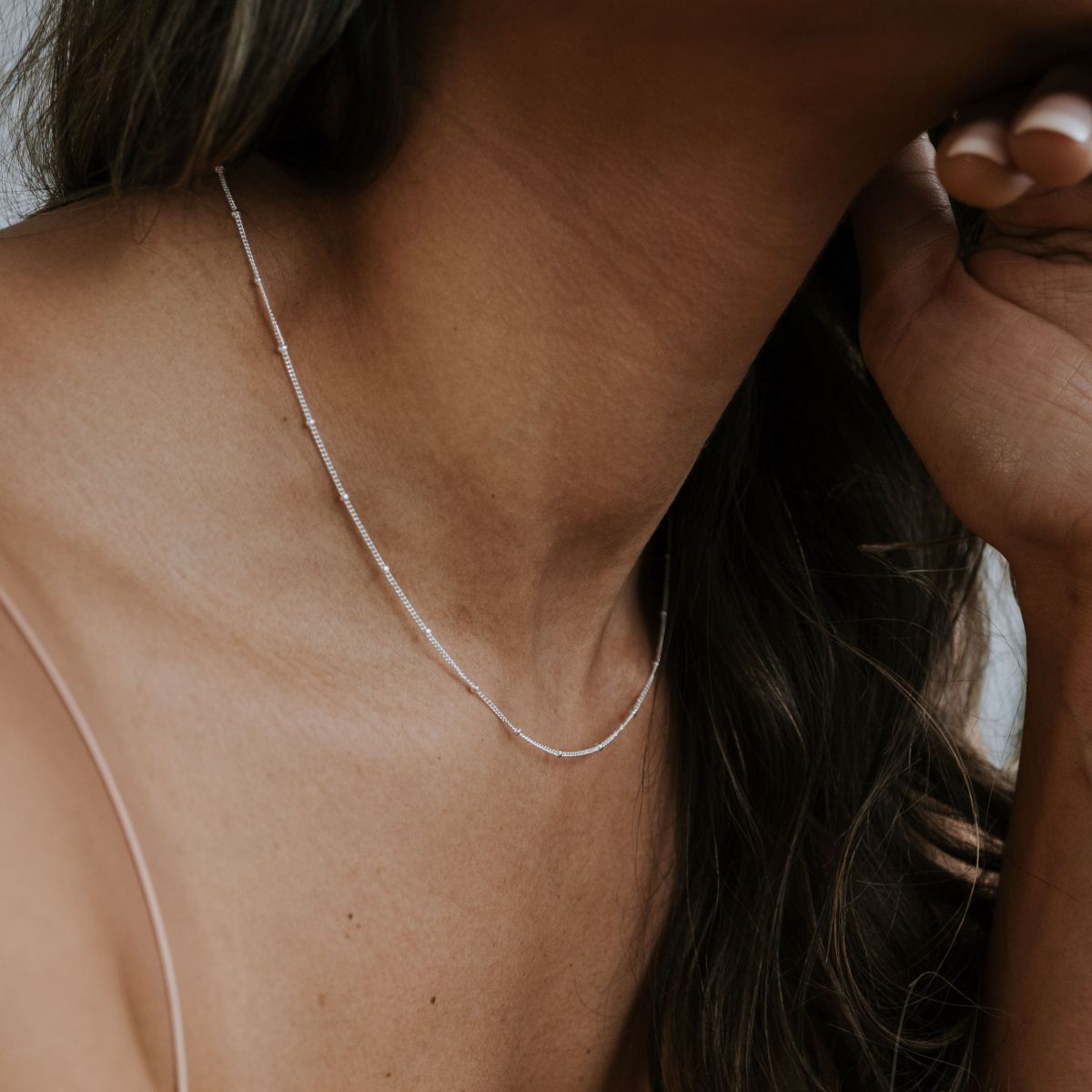 A woman wears a Becoming Jewelry Satellite Chain Necklace, featuring a delicate, thin silver chain with small beads, against her bare shoulder. She tilts her head slightly while resting her hand near her face.
