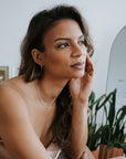 A woman with long hair, seated indoors near plants and a mirror, rests her head on her hand. She wears the Becoming Jewelry Satellite Chain Necklace, which adds elegance to her contemplative gaze.