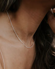 A woman wears a Becoming Jewelry Satellite Chain Necklace, featuring a delicate, thin silver chain with small beads, against her bare shoulder. She tilts her head slightly while resting her hand near her face.