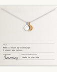 The "Count My Blessings Necklace" by Becoming Jewelry features a silver necklace with a round and crescent moon pendant, elegantly displayed on paper inscribed with the thoughtful phrase, "When I count my blessings, I count you twice," alongside other publication details. This charming gift pairs beautifully with silver drop discs.