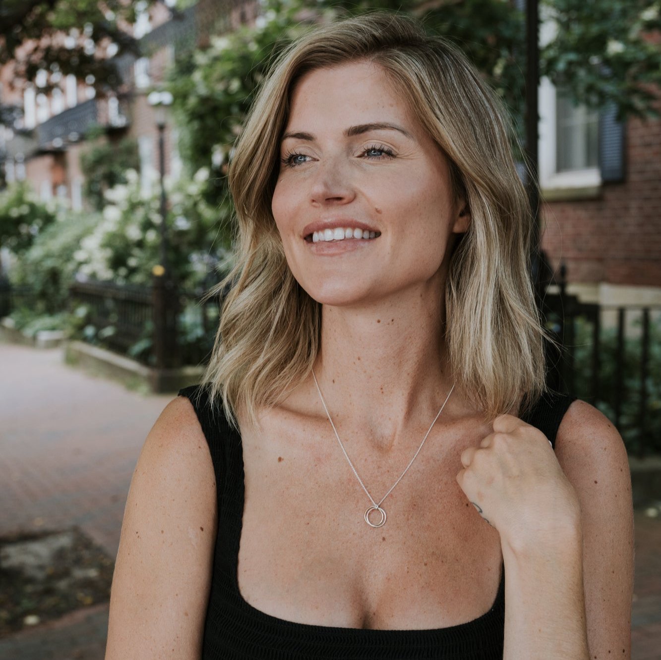 A woman with shoulder-length blonde hair, wearing a black top and a Friendship Circles Necklace by Becoming Jewelry, is smiling outdoors in a garden setting.