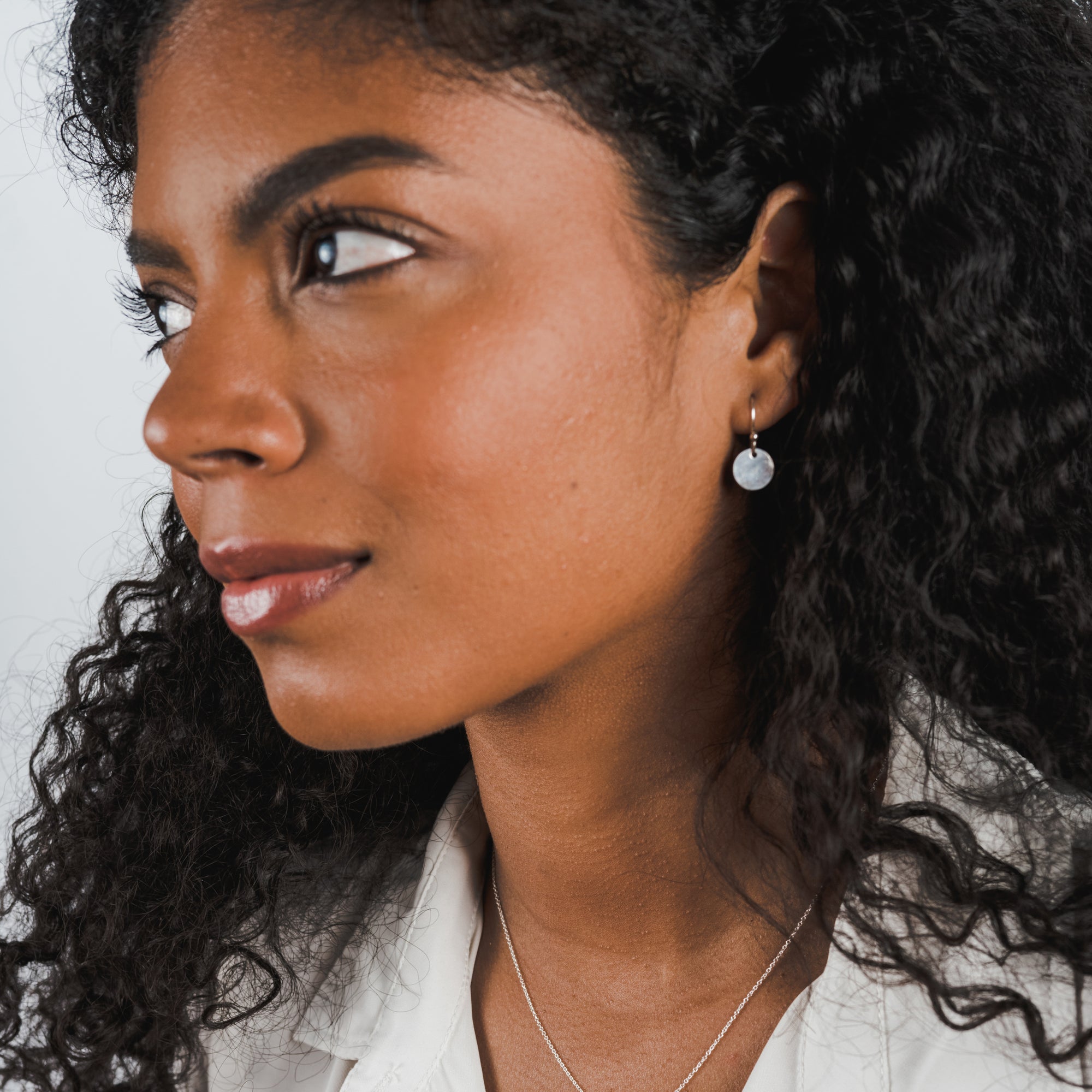 A person with curly hair and wearing lightweight tiny Hammered Disc Drop Earrings by Becoming Jewelry looks to the side. They are also wearing a white top and a hammered-by-hand necklace.