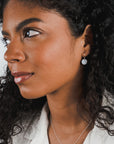 A person with curly hair and wearing lightweight tiny Hammered Disc Drop Earrings by Becoming Jewelry looks to the side. They are also wearing a white top and a hammered-by-hand necklace.