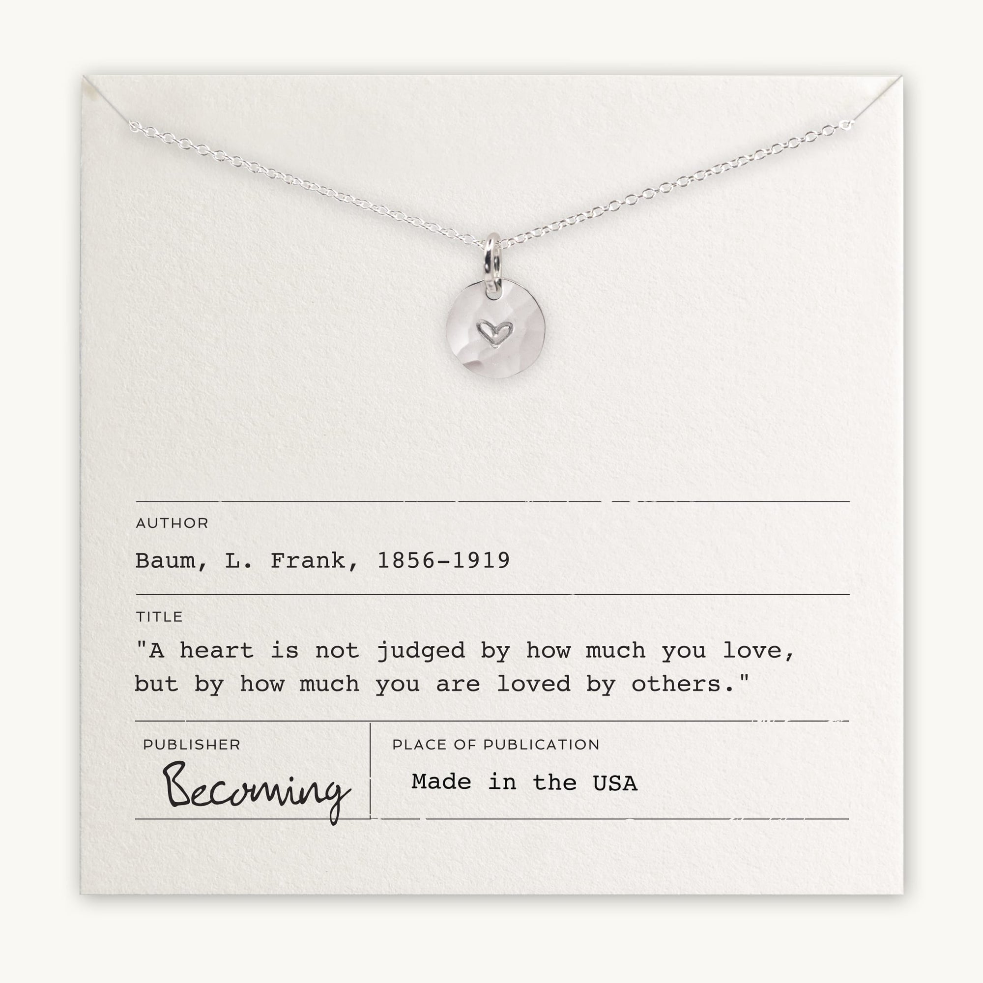 The Heart Necklace by Becoming Jewelry is a silver necklace with a round pendant featuring a small heart symbol. It comes placed on a card that quotes L. Frank Baum: "A heart is not judged by how much you love, but by how much you are loved by others." This meaningful gift captures the essence of true affection.