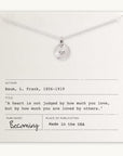 The Heart Necklace by Becoming Jewelry is a silver necklace with a round pendant featuring a small heart symbol. It comes placed on a card that quotes L. Frank Baum: "A heart is not judged by how much you love, but by how much you are loved by others." This meaningful gift captures the essence of true affection.