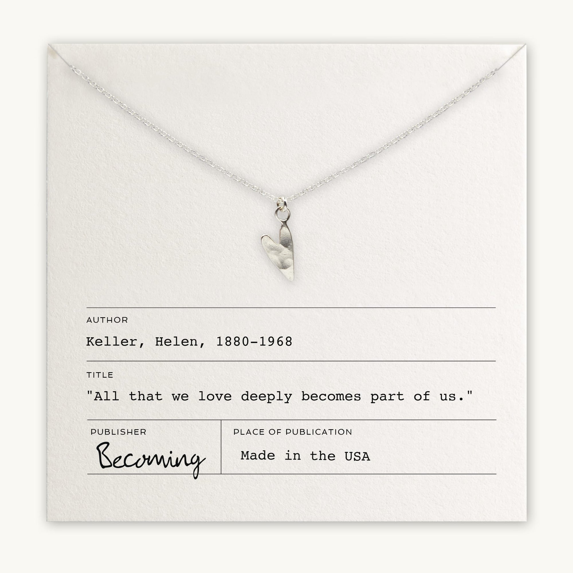 A silver heart charm pendant on the Love Deeply Necklace by Becoming Jewelry is placed over a card featuring the quote by Helen Keller, "All that we love deeply becomes part of us." The card includes the author's name, title, and publication details.
