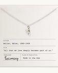 A silver heart charm pendant on the Love Deeply Necklace by Becoming Jewelry is placed over a card featuring the quote by Helen Keller, "All that we love deeply becomes part of us." The card includes the author's name, title, and publication details.