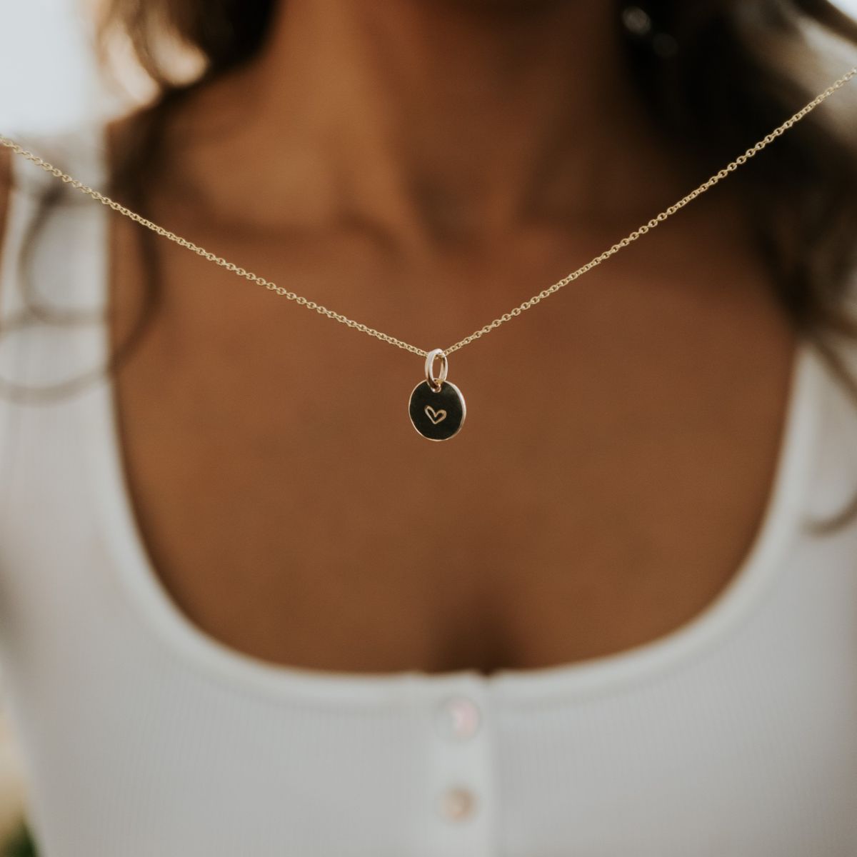 A person wears the Becoming Jewelry Heart Necklace, with its gold round pendant featuring a tiny heart engraving, against a white button-up top—an exquisite gift cherished by others.