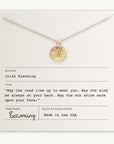 An Irish Blessing Necklace from Becoming Jewelry, featuring a gold pendant, rests elegantly on a card titled "Irish Blessing" adorned with a heartwarming quote. Made in the USA and perfect for gifting to those embarking on new adventures, this necklace is an ideal way to offer well-wishes.