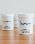 Three white containers labeled "Becoming Jewelry Cleaner" sit on a wooden surface. Inside, the liquid cleaner provides an ideal solution for your precious pieces, with each container equipped with a convenient dip tray to ensure every gem sparkles like new.