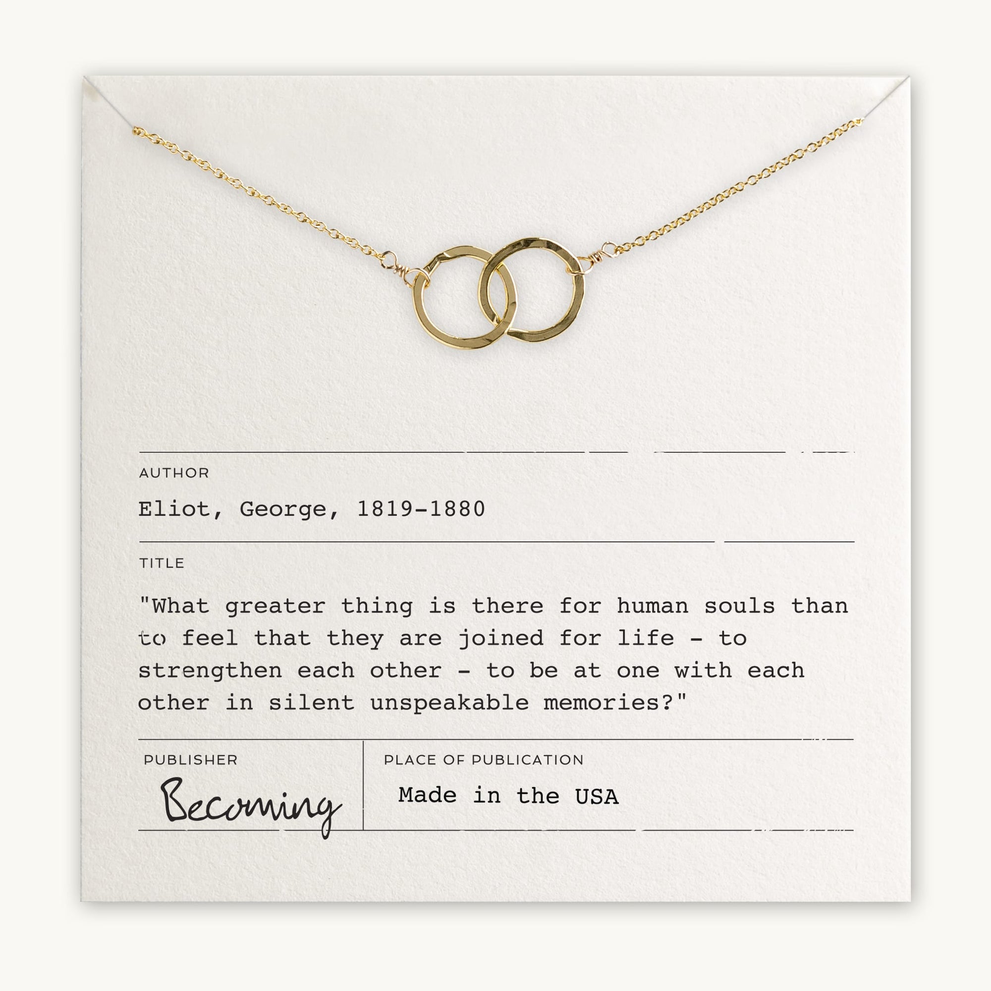 A gold "Joined for Life" necklace with two intertwined circles rests on a note. The note displays a quote by George Eliot from the book "Becoming Jewelry." It mentions that the necklace is made in the USA.