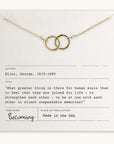 A gold "Joined for Life" necklace with two intertwined circles rests on a note. The note displays a quote by George Eliot from the book "Becoming Jewelry." It mentions that the necklace is made in the USA.