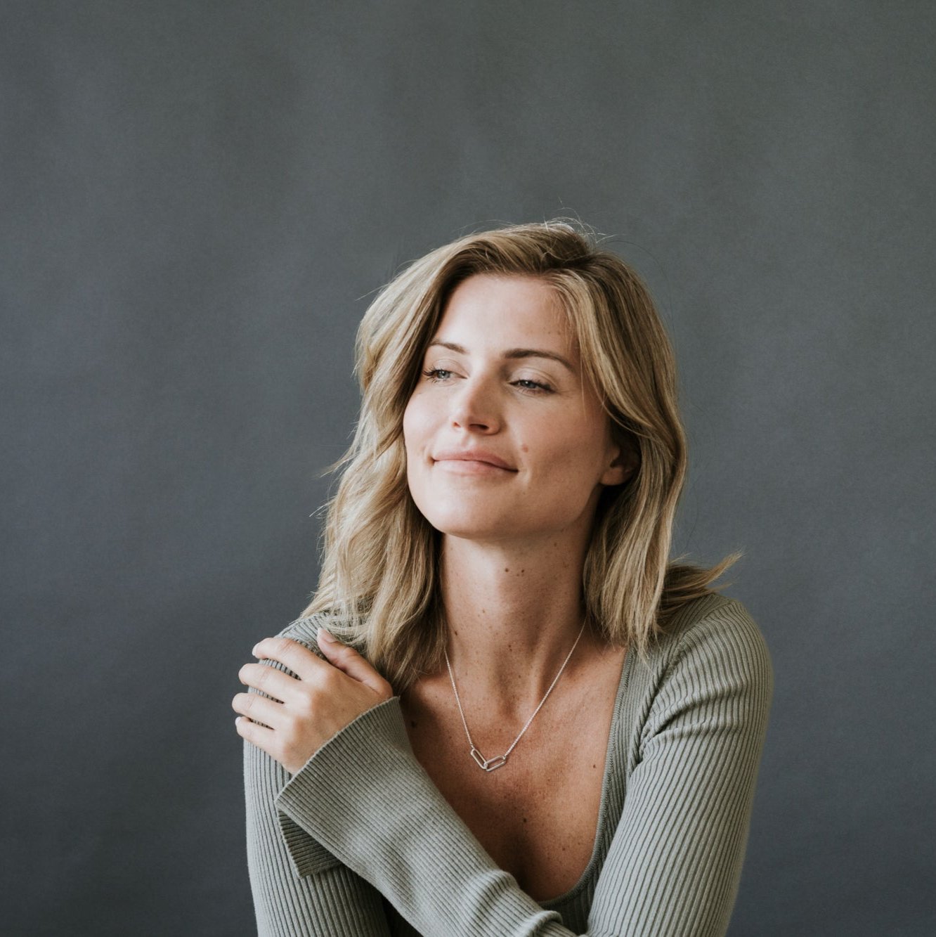 A person with shoulder-length blonde hair, wearing a light green sweater and a Linked Together Necklace from Becoming Jewelry, sits against a plain dark gray background. They have a slight smile and one hand resting on their shoulder, cherishing the special connection this gift represents.