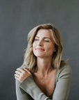 A person with shoulder-length blonde hair, wearing a light green sweater and a Linked Together Necklace from Becoming Jewelry, sits against a plain dark gray background. They have a slight smile and one hand resting on their shoulder, cherishing the special connection this gift represents.