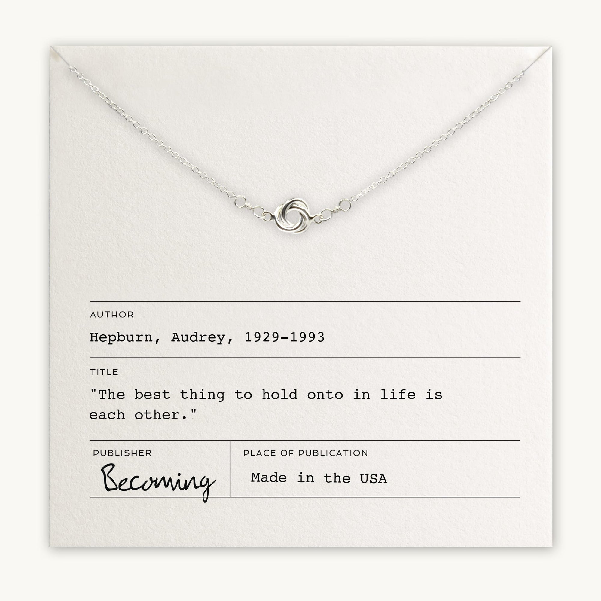 A delicate silver Love Knot necklace from Becoming Jewelry, featuring an intertwined ring pendant, is displayed on a card quoting Audrey Hepburn: "The best thing to hold onto in life is each other." This heartfelt gift, featuring a small knot charm, is proudly made in the USA.