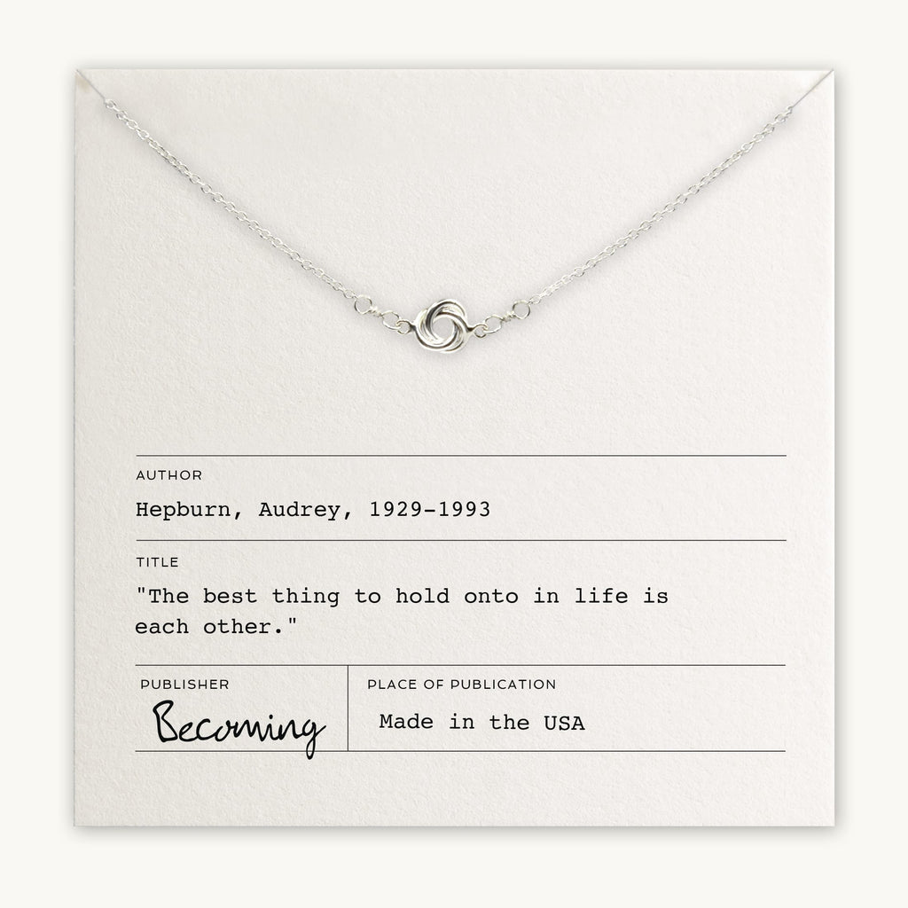 A Love Knot Necklace laid over a card with a quote by Audrey Hepburn, titled "Becoming," made in the USA by Becoming Jewelry.