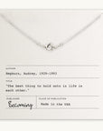 A delicate silver Love Knot necklace from Becoming Jewelry, featuring an intertwined ring pendant, is displayed on a card quoting Audrey Hepburn: "The best thing to hold onto in life is each other." This heartfelt gift, featuring a small knot charm, is proudly made in the USA.