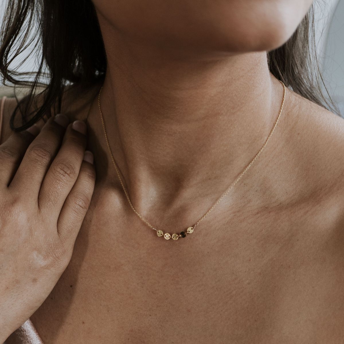 A close-up of the Becoming Jewelry Mothers & Daughters Necklace reveals its elegant gold design with small circular pendants, captured against a softly blurred background. Resting by her neck, it symbolizes the timeless bond between strong mothers and daughters.