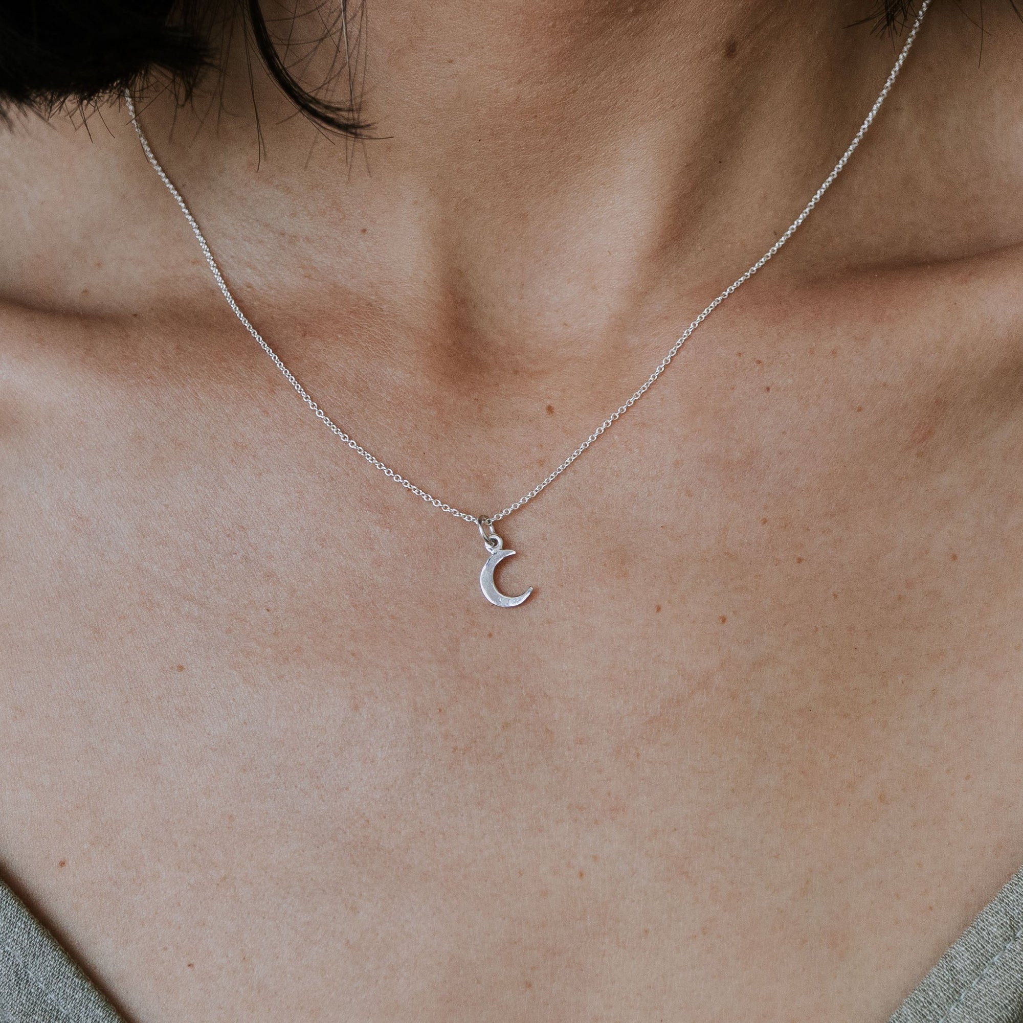 Close-up of a person wearing a thin silver chain necklace with a crescent moon pendant—an elegant and meaningful piece known as the Love You To The Moon Necklace by Becoming Jewelry. The person is dressed in a light-colored top, making the necklace stand out beautifully.