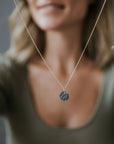 A remarkable woman holds a silver River Necklace from Becoming Jewelry, featuring a circular pendant adorned with a leaf imprint design that echoes her indomitable spirit.