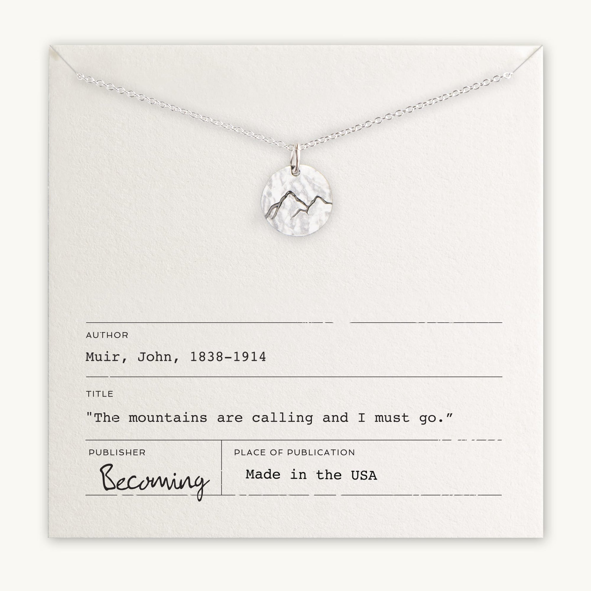 The "Mountains Are Calling Necklace" by Becoming Jewelry showcases a silver necklace adorned with a small mountain charm pendant, gracefully presented on a card. The card features an inspiring quote by John Muir: "The mountains are calling and I must go." It's the ideal gift for nature and adventure enthusiasts.