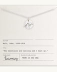 The "Mountains Are Calling Necklace" by Becoming Jewelry showcases a silver necklace adorned with a small mountain charm pendant, gracefully presented on a card. The card features an inspiring quote by John Muir: "The mountains are calling and I must go." It's the ideal gift for nature and adventure enthusiasts.