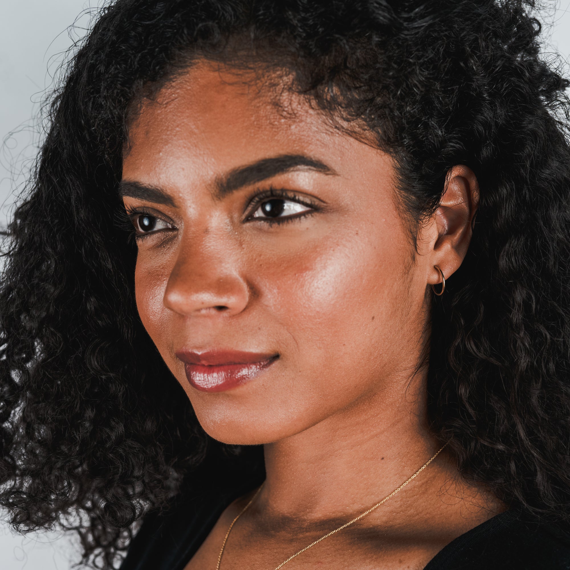 A woman with curly dark hair, wearing medium open hoop earrings from Becoming Jewelry and a gold necklace, gazes slightly off-camera with a neutral expression. She has a glowing complexion and is dressed in a classic black top.