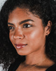 A woman with curly dark hair, wearing medium open hoop earrings from Becoming Jewelry and a gold necklace, gazes slightly off-camera with a neutral expression. She has a glowing complexion and is dressed in a classic black top.