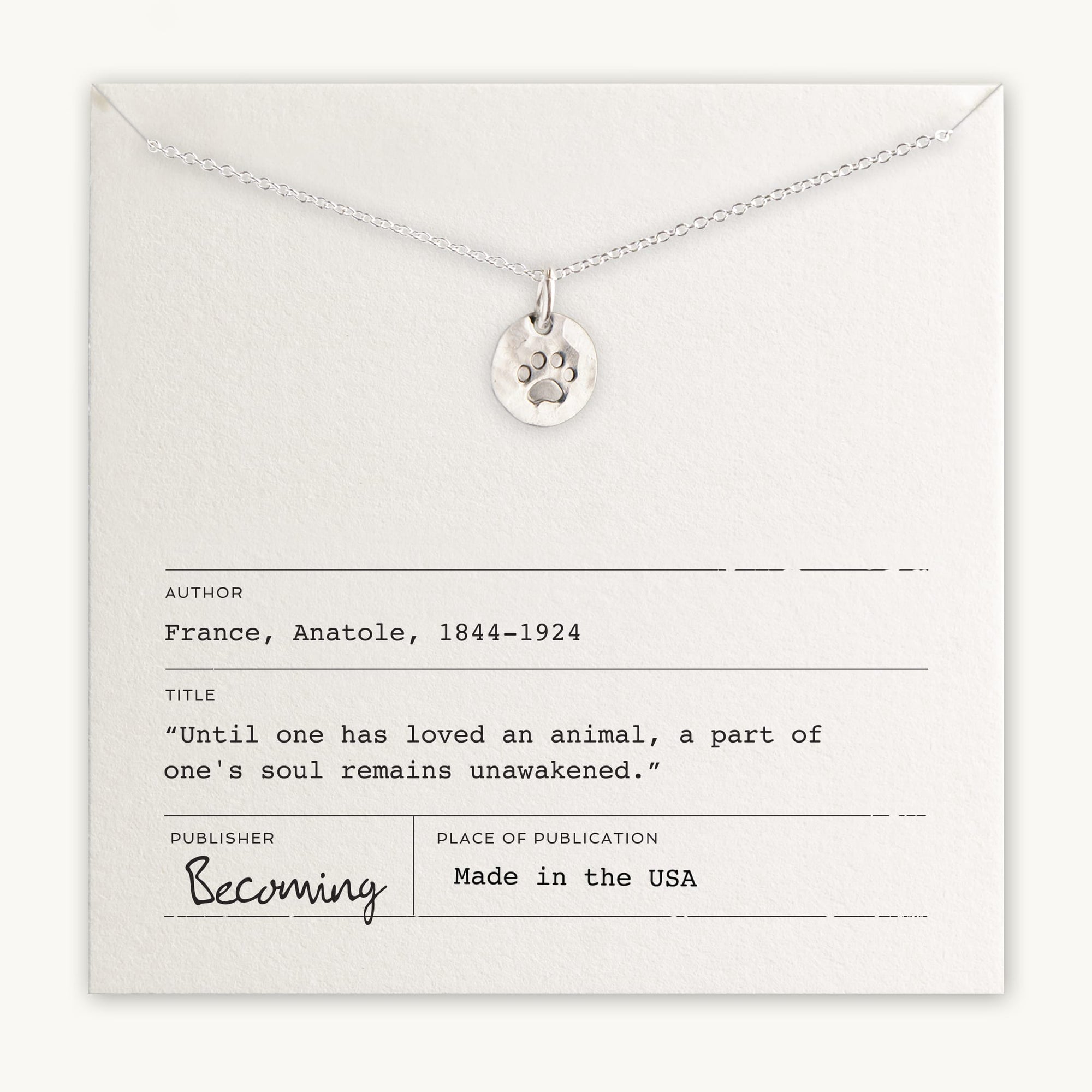 The Paw Print Necklace by Becoming Jewelry is elegantly displayed on an off-white card that includes a quote from Anatole France about loving animals, details about the author, and proudly states "Made in the USA." This ideal gift for animal lovers perfectly captures the essence of adoration for our furry friends.