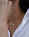 A person wearing a white shirt is shown with a focus on the "Simple Pleasures Pearl Necklace" by Becoming Jewelry, featuring a single pearl pendant around their neck, exuding timeless beauty.