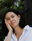 A woman with shoulder-length dark hair and a white shirt rests her head on her hand while smiling, adorned with Becoming Jewelry's Simple Pleasures Pearl Necklace, set against a green, blurred background.