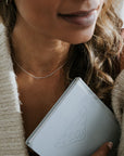A person clutches a blue book close to their heart, wearing a knitted sweater and the Path Necklace by Becoming Jewelry, with long, wavy hair partially visible.