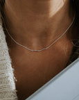 Close-up of a person wearing the Becoming Jewelry Path Necklace, a silver piece with a heart-like curve design, paired with a knitted sweater while holding a book.