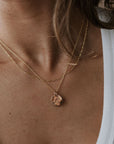 A person in a white top wears two gold necklaces; one is a simple chain, and the River Necklace by Becoming Jewelry boasts a round pendant, reflecting the indomitable spirit of remarkable women.