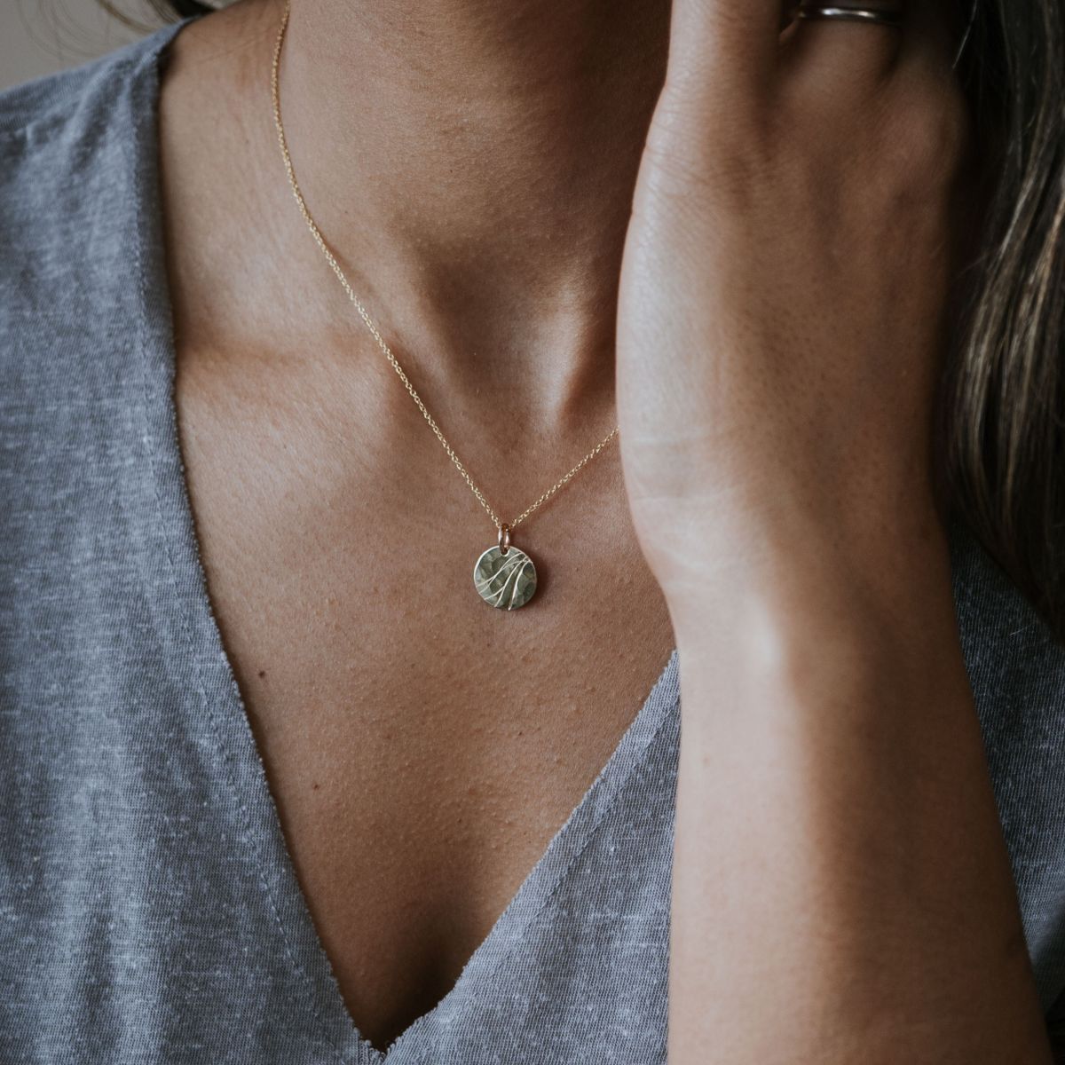 A person in a gray V-neck shirt wears the stunning River Necklace by Becoming Jewelry, featuring a round gold pendant. Their hand partially covers their face, showcasing an indomitable spirit.