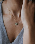 A person in a gray V-neck shirt wears the stunning River Necklace by Becoming Jewelry, featuring a round gold pendant. Their hand partially covers their face, showcasing an indomitable spirit.