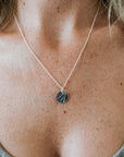 A close-up view of a person wearing a silver River Necklace from Becoming Jewelry. The necklace features a round, dark pendant with an intricate silver branch design. The person is dressed in a green top, embodying the indomitable spirit of remarkable women.