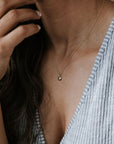 A person in a striped shirt wears the Shine Necklace by Becoming Jewelry, featuring a delicate pendant that could be cubic zirconia, and raises their hand near their face.